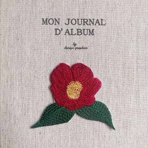 Pode incluir: Um diário com o texto "MON JOURNAL D'ALBUM" e "by design graphics" bordado numa capa com textura de linho. Uma flor vermelha com centro amarelo e folhas verdes é bordada na capa.