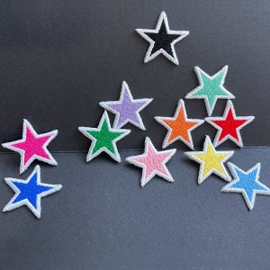 Pode incluir: Uma coleção de patches bordados em forma de estrela em várias cores, incluindo rosa, azul, verde e amarelo, cada um com uma borda branca. Os patches estão espalhados sobre uma superfície cinza escuro.