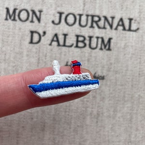 May include: Embroidered patch of a cruise ship with a white body, blue hull, and a red and blue smokestack. The patch is small and detailed, perfect for adding to clothing or accessories. The background has the text "MON JOURNAL D'ALBUM".