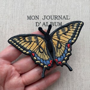 May include: Embroidered butterfly patch with black, yellow, blue, and red details. The butterfly has detailed wing patterns and is set against a background with the text "MON JOURNAL D'ALBUM".