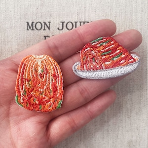Może przedstawiać: Dwie haftowane naszywki przedstawiające kimchi. Jedna przedstawia pojedynczy kawałek, druga stos na talerzu. Naszywki są pomarańczowe, czerwone, zielone i białe, ze szczegółowymi przeszyciami. W tle napis "MON JOUR".
