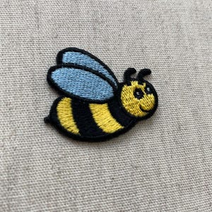 May include: Embroidered bee patch with yellow and black stripes, blue wings, and a smiling face. The patch is outlined in black and is designed to be sewn onto fabric. The bee is a cute, cartoon-style design.