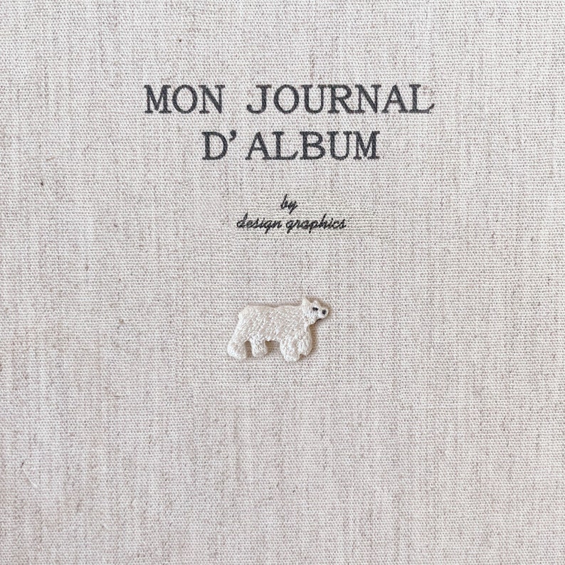K&ouml;nnte beinhalten: Ein beiger Stoff-Journal-Einband mit dem Text "MON JOURNAL D'ALBUM" und "by design graphics" in Schwarz. Ein kleiner, wei&szlig; gestickter Eisb&auml;r ist unter dem Text zentriert.