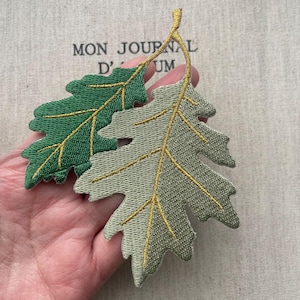 以下が含まれることがあります： 刺繍された2つの葉のパッチ。1つは鮮やかな緑色、もう1つはくすんだセージグリーンです。どちらも金色の葉脈と茎が特徴です。パッチは手で持たれており、背景には「MON JOURNAL D'UM」の文字が見えます。