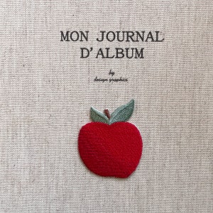 Puede incluir: Una cubierta de diario de tela beige con el texto "MON JOURNAL D'ALBUM" y "by design graphics" impreso en negro. Una manzana roja con hojas verdes está bordada en la cubierta.