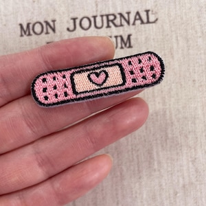 Può includere: Una toppa ricamata a forma di cerotto adesivo, caratterizzata da un design rettangolare rosa con un bordo nero cucito. Il centro della toppa presenta un quadrato color pesca chiaro con una piccola icona a forma di cuore rosa scuro.