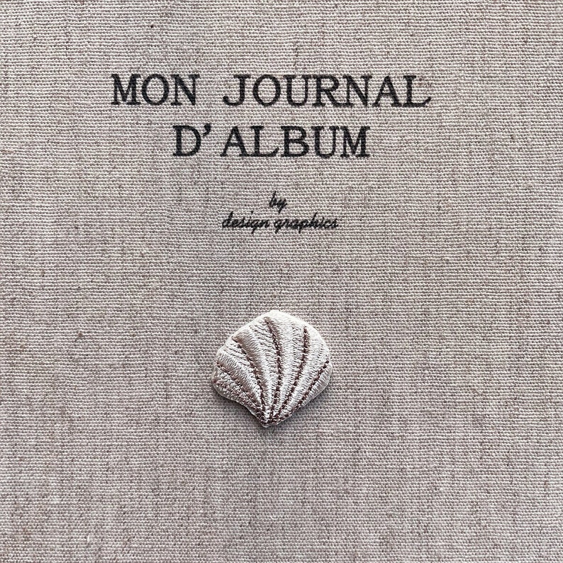 K&ouml;nnte beinhalten: Nahaufnahme eines beigefarbenen Stoffes mit dem Text "MON JOURNAL D'ALBUM" und "by design graphics" in Schwarz. Eine kleine, gestickte Muschel ist unten zentriert. Der Stoff hat ein strukturiertes Aussehen.
