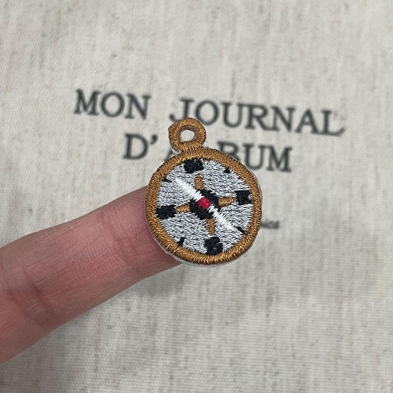May include: Embroidered compass patch with a gold border, black and red accents, and a silver-grey face. The patch is held up by a finger. The text "MON JOURNAL D'RUM" is in the background.