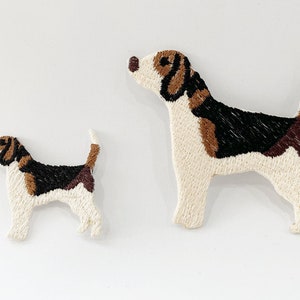 May include: Two embroidered dog figures, one large and one small, against a white backdrop. The dogs are white with brown, black, and tan markings. The larger dog is standing, looking upwards, and the smaller dog is in a similar pose.