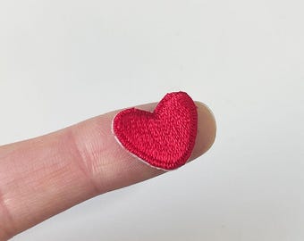 May include: A small, embroidered red heart patch with a white border. The heart is resting on a finger, showing the patch's size and detail. The patch is likely for clothing or craft projects.