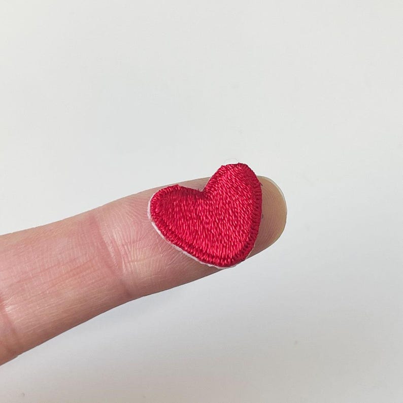 K&ouml;nnte beinhalten: Ein kleiner, gestickter roter Herzaufn&auml;her mit wei&szlig;em Rand. Das Herz liegt auf einem Finger und zeigt die Gr&ouml;&szlig;e und die Details des Aufn&auml;hers. Der Aufn&auml;her ist wahrscheinlich f&uuml;r Kleidung oder Bastelprojekte gedacht.
