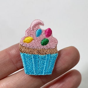 May include: Embroidered cupcake patch. The cupcake has light pink icing, a brown base, and a blue case. It's decorated with colourful sprinkles. Ideal for adding to clothes or accessories. The patch measures approximately 3 cm in height.
