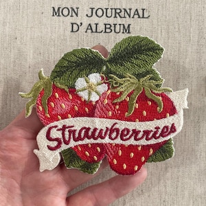 Puede incluir: Parche bordado con dos fresas rojas con hojas verdes y una flor blanca. Una pancarta sobre las fresas dice "Strawberries" en hilo burdeos. El fondo tiene el texto "MON JOURNAL D'ALBUM".