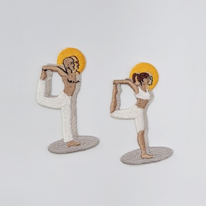 May include: Two embroidered patches depicting figures in yoga poses. The figures are in white and tan, with a yellow halo. They are standing on a gray oval base. The patches are on a white background.