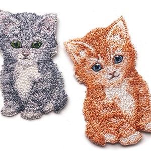 Könnte beinhalten: Zwei gestickte Katzenflicken. Einer ist grau und weiß mit grünen Augen, der andere ist orange und weiß mit blauen Augen. Beide Flicken haben eine flauschige Textur und sind vor einem weißen Hintergrund platziert.