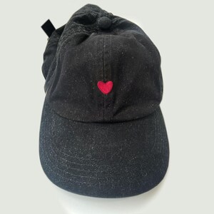 K&ouml;nnte beinhalten: Schwarze Baseballkappe mit einem kleinen, gestickten roten Herz auf der Vorderseite. Die Kappe hat einen gebogenen Schirm und einen Stoffriemen mit Metallschnalle hinten. Hergestellt aus einem weichen, strukturierten Material.