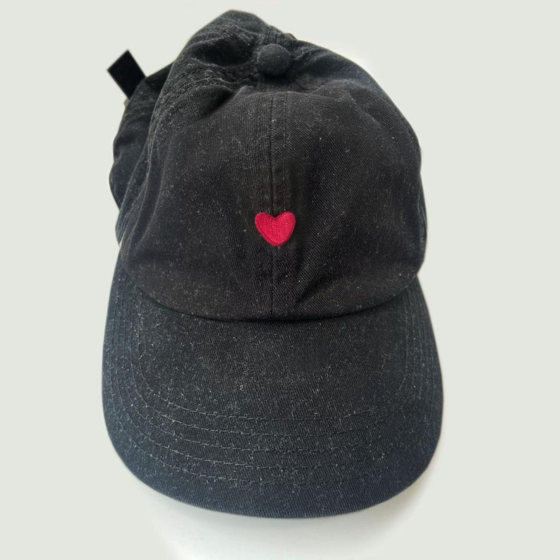K&ouml;nnte beinhalten: Schwarze Baseballkappe mit einem kleinen, gestickten roten Herz auf der Vorderseite. Die Kappe hat einen gebogenen Schirm und einen Stoffriemen mit Metallschnalle hinten. Hergestellt aus einem weichen, strukturierten Material.