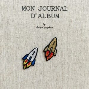 Może przedstawiać: Dwie haftowane naszywki w kształcie rakiety. Jedna jest biała z niebieskimi i żółtymi płomieniami, druga jest złota z czerwonymi i żółtymi płomieniami. Tło to beżowa tkanina z napisem "MON JOURNAL D'ALBUM" i "by design graphics".
