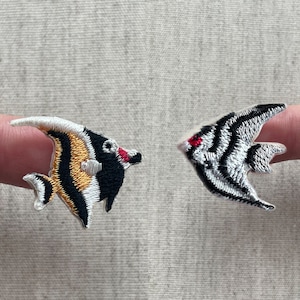 May include: Two embroidered fish patches. One patch features a black, white, and gold fish with a red accent. The other patch is a black and white striped fish with a red accent. Both patches have a detailed design.