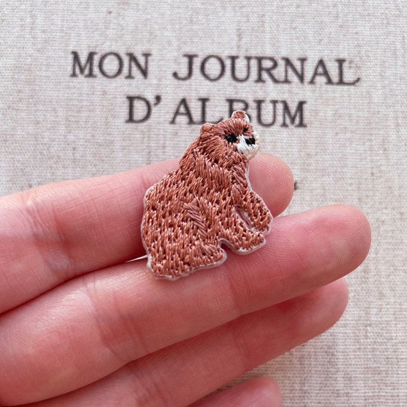 May include: Embroidered brown bear patch with black and white details. The bear is sitting, with a detailed stitched texture. The background includes the text "MON JOURNAL D'ALBUM".