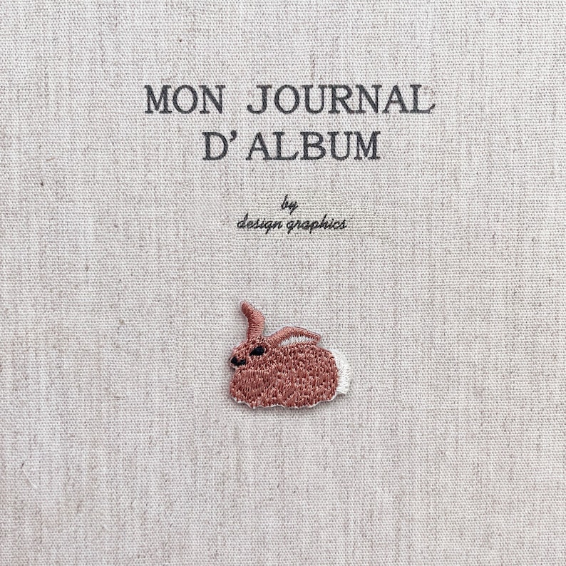 K&ouml;nnte beinhalten: Ein beiger Stoff-Journal-Einband mit dem Text "MON JOURNAL D'ALBUM" und "by design graphics" in Schwarz. Auf dem Einband befindet sich ein kleiner, gestickter braun-wei&szlig;er Hase.