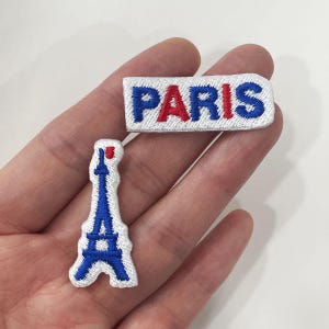 May include: Two embroidered patches: one rectangular with "PARIS" in blue and red, and another shaped like the Eiffel Tower in blue. Both patches have a white background and are likely for clothing or accessories.