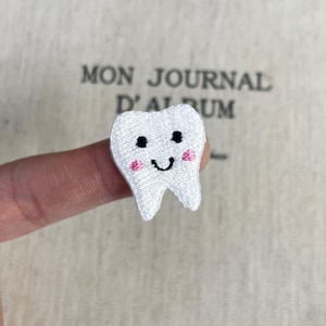 May include: A small white embroidered patch shaped like a tooth with a smiling face. The tooth features two black circular eyes, a curved black mouth, and two small pink circular cheeks. The embroidery has a textured, thread-like appearance.