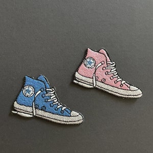 May include: Embroidered patches of high-top sneakers, one blue and one pink, with white laces and a circular logo on the side. The patches have a gray border and are set against a dark gray background.