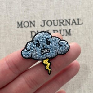 Puede incluir: Parche bordado con una nube de dibujos animados con una cara, en tonos azules y grises, con un rayo amarillo. El parche tiene un borde negro y probablemente sea para ropa o accesorios.