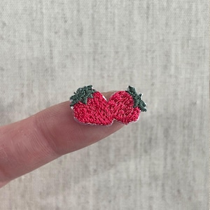 Puede incluir: Un pequeño parche bordado con dos fresas rojas con copas verdes. Las fresas están cosidas con una textura detallada y se muestran en un dedo. El parche es probablemente para ropa o proyectos de artesanía.