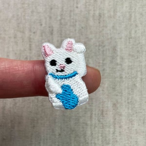 Könnte beinhalten: Gestickter Aufnäher eines weißen Cartoon-Katze mit rosa Ohren und einem blauen Schal. Die Katze hält ein blaues Objekt. Der Aufnäher ist klein und detailliert, mit schwarzen Augen und einem lächelnden Mund. Für Kleidung oder Accessoires.