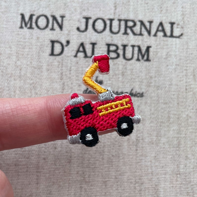 May include: A small, embroidered patch of a red fire truck with yellow and silver accents. The fire truck has black wheels and a ladder. The patch is held up by a finger. The background has the text "MON JOURNAL D'ALBUM".