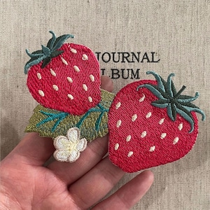 May include: Embroidered strawberry patches with red fruit, white seeds, and green leaves. The patches have a detailed design and are likely for decorative purposes. The background has the text "JOURNAL BUM".