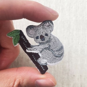 May include: Embroidered patch featuring a gray koala bear clinging to a brown tree branch with a green leaf. The koala has detailed stitching, with a focus on its fur and facial features. The patch is held by a hand.