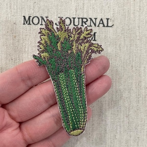 May include: Embroidered celery patch with green and purple details. The patch is held in a hand, with the text "MON JOURNAL" visible in the background.