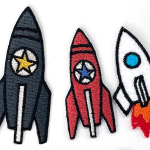 May include: Three embroidered rocket ship patches in black, red, and white. Each rocket has a star emblem in the centre. The black rocket has a yellow star, the red has a blue star, and the white rocket has a blue circle with orange flames.