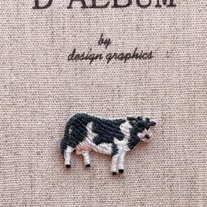 Könnte beinhalten: Gestickter Aufnäher einer schwarz-weißen Kuh mit rosa Akzenten. Der Aufnäher befindet sich auf einem beigefarbenen Stoffhintergrund mit dem Text "D'Album by design graphics". Die Kuh steht und hat detaillierte Stickereien.