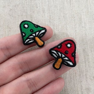 Puede incluir: Dos parches bordados con forma de seta. Uno es verde con puntos blancos, el otro es rojo con puntos blancos. Ambos tienen contornos negros, branquias blancas y tallos naranjas. Estos parches son probablemente para ropa o accesorios.