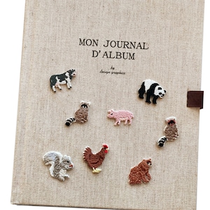 May include: A beige journal cover with embroidered animal patches, including a cow, panda, raccoon, pig, squirrel, chicken, and bear. The journal cover has the text "MON JOURNAL D'ALBUM" and "by design graphics".
