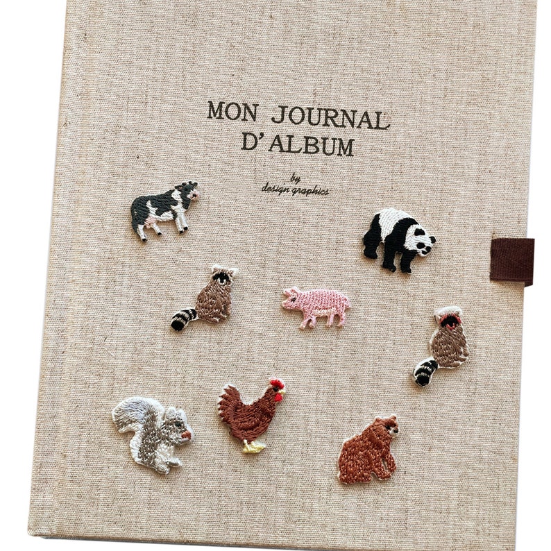 May include: A beige journal cover with embroidered animal patches, including a cow, panda, raccoon, pig, squirrel, chicken, and bear. The journal cover has the text "MON JOURNAL D'ALBUM" and "by design graphics".