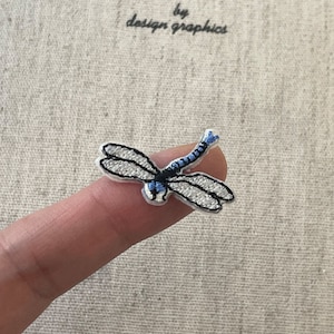 May include: Embroidered dragonfly patch with white wings, black outlines, and a blue body. The patch is small and detailed, with the text "by design graphics" in the upper left corner.