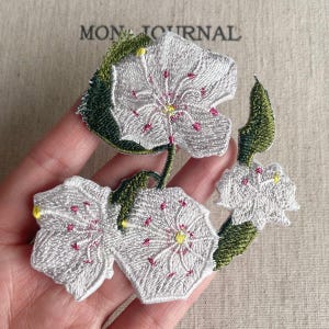 May include: Embroidered floral patch featuring three white flowers with pink accents and yellow centers, accompanied by green leaves. The patch is intricately detailed and appears to be hand-stitched. The text "MON JOURNAL" is visible in the background.