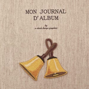 Könnte beinhalten: Ein beigefarbener Stoffeinband für ein Journal mit dem gestickten Text MON JOURNAL D'ALBUM by o-check design graphics. Unter dem Text sind zwei goldgelbe Glocken mit braunen Griffen in gekreuzter Position auf das strukturierte Material gestickt.