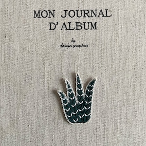 May include: A journal cover with the text "MON JOURNAL D'ALBUM" and "by design graphics" in black. A dark green embroidered plant design with white stitching is centered at the bottom. The background is a light beige fabric.