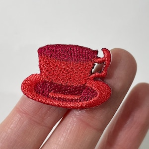 May include: Embroidered patch of a red teacup and saucer. The teacup has a handle and is a darker shade of red at the top. The saucer is a lighter shade of red. The patch is likely for clothing or accessories.