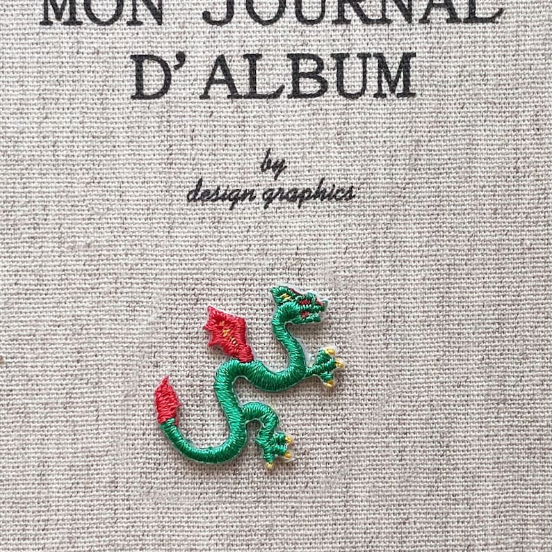 May include: Embroidered green dragon patch with red accents, featuring the text "MON JOURNAL D'ALBUM" and "by design graphics" on a textured, light-colored fabric. The dragon has yellow claws and a red tail.