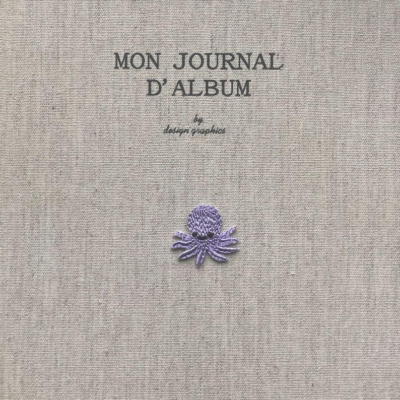 Pu&ograve; includere: Una copertina di album in tessuto grigio chiaro con la scritta "MON JOURNAL D'ALBUM" e "by design graphics" stampata in nero. Un piccolo polpo viola ricamato &egrave; centrato in basso. La copertina dell'album ha un aspetto strutturato.