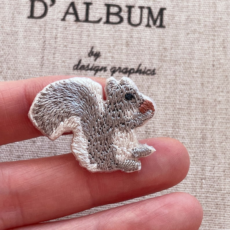 May include: Embroidered patch featuring a gray and white squirrel with a fluffy tail and a reddish-brown nose. The patch is in the shape of a squirrel and has a white border. The text "D'ALBUM by design graphics" is in the background.