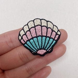 Peut inclure: Écusson brodé en forme de coquillage avec une bordure noire. La coquille présente des sections beige clair, rose et bleu turquoise. L'écusson est un article décoratif, probablement pour les vêtements ou les accessoires.