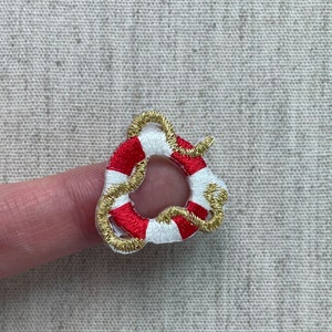 May include: Embroidered miniature life preserver with red and white stripes, accented with gold thread. The small, circular design is a nautical-themed craft element, perfect for embellishing clothing or accessories.